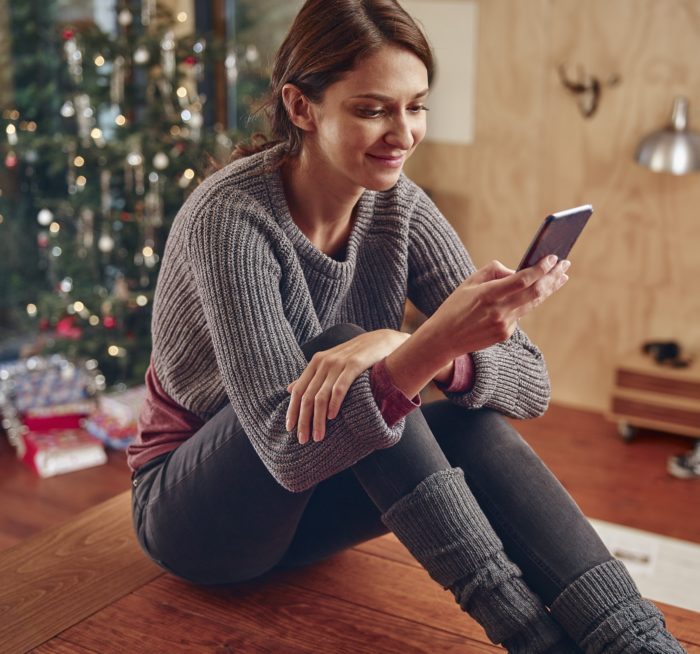 Young woman sitting on flooor in front of Christmas tree using smart phone